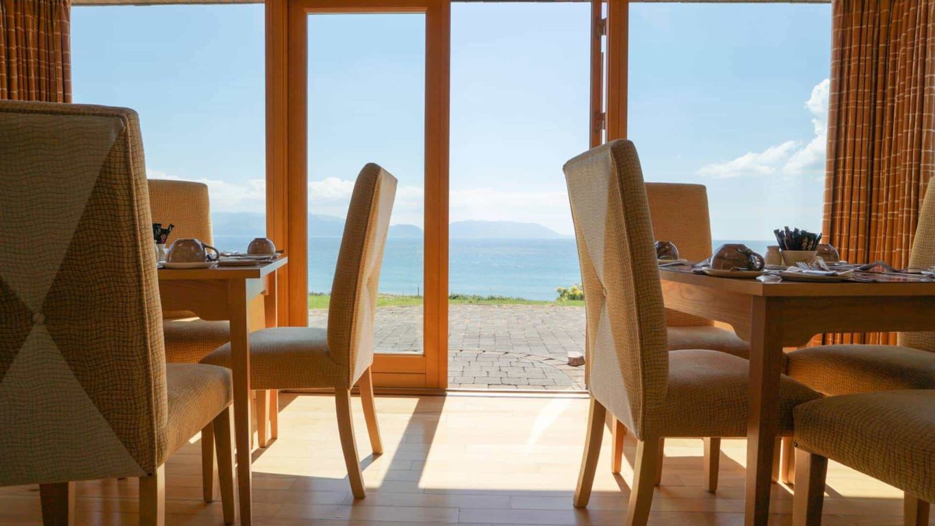 Sunlit dining area with cosy seating, offering stunning sea views through large windows, perfect for a relaxing meal.