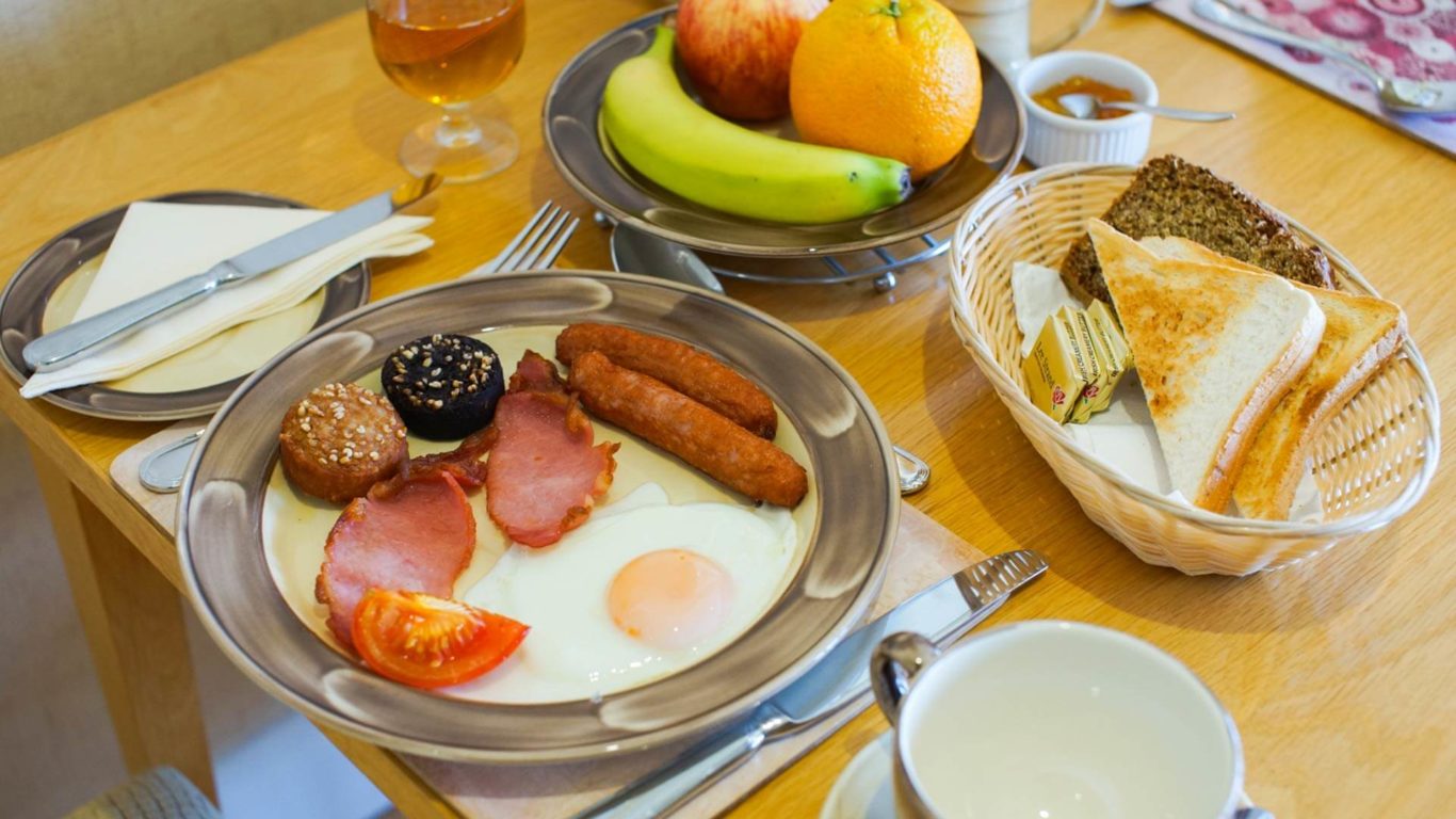 Enjoy a full English breakfast with toast, sausages, eggs, and fresh fruit, set on a cosy hotel dining table.