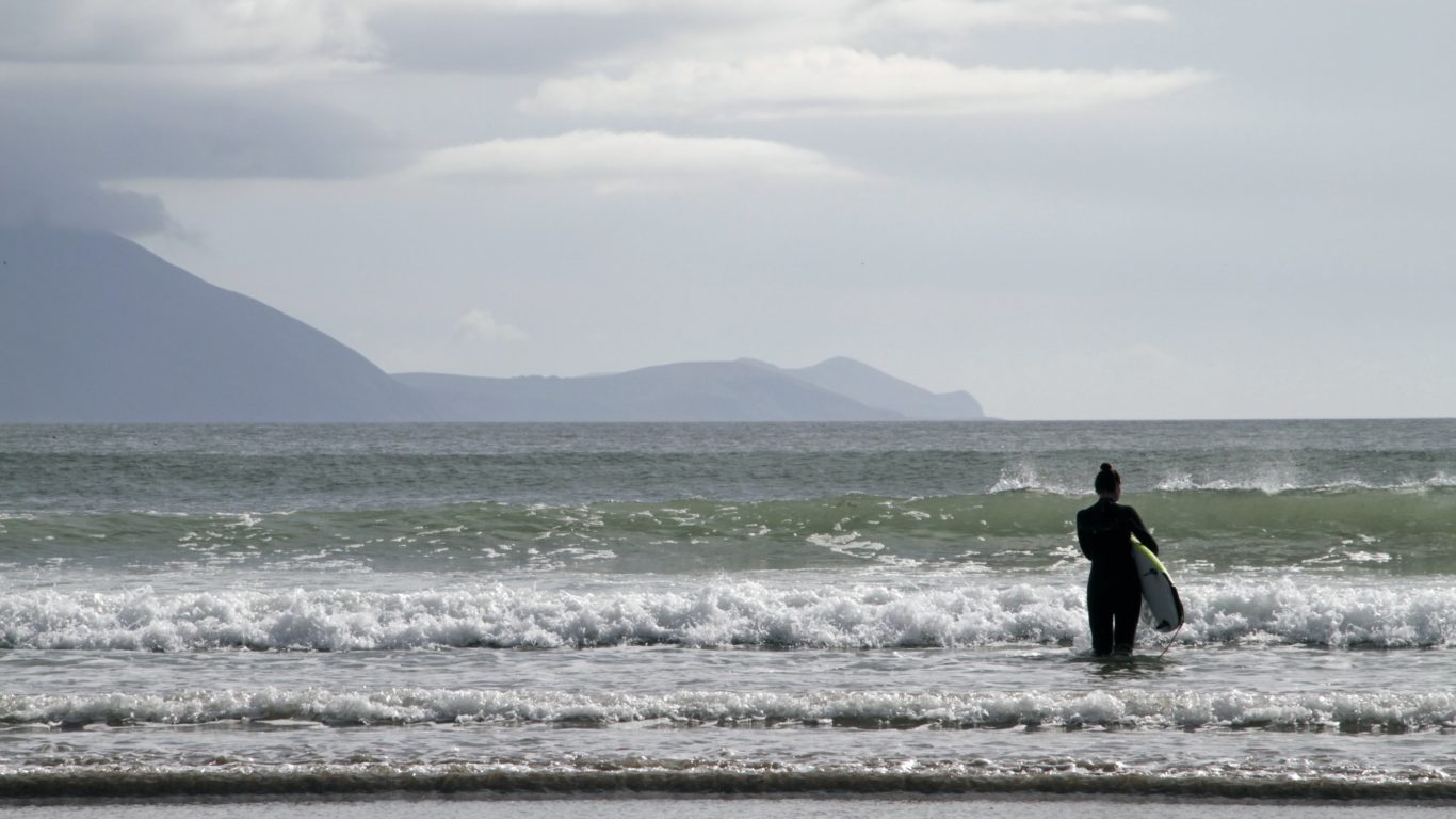 woman-with-a-surfing-board-in-the-sea-surrounded-by-the-hills-under-a-cloudy-sky-in-ireland-171718954