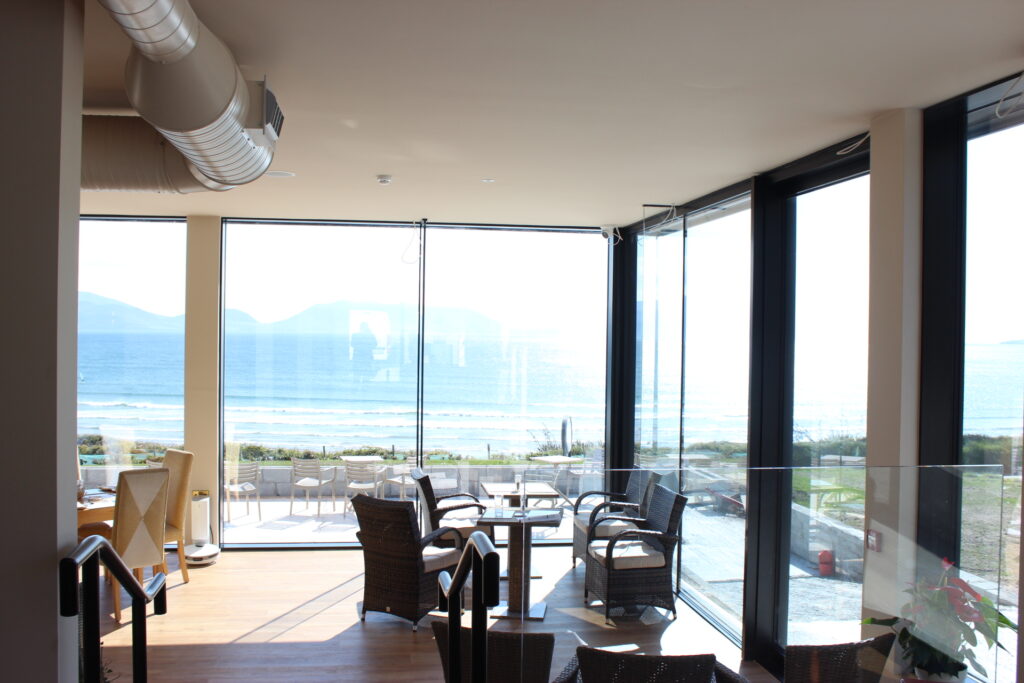 Indoor seating area with wicker chairs and glass tables, large floor-to-ceiling windows offering a view of the beach and ocean outside.
