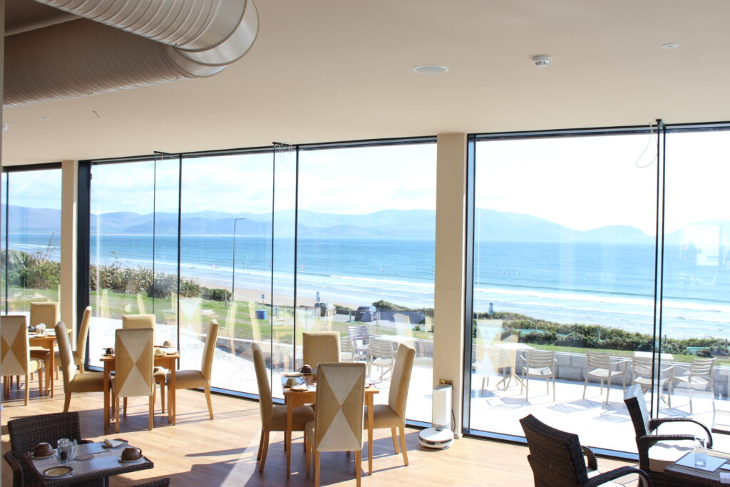 Bright restaurant dining area with tables and chairs arranged by large glass windows overlooking a sandy beach and ocean with mountains in the distance.