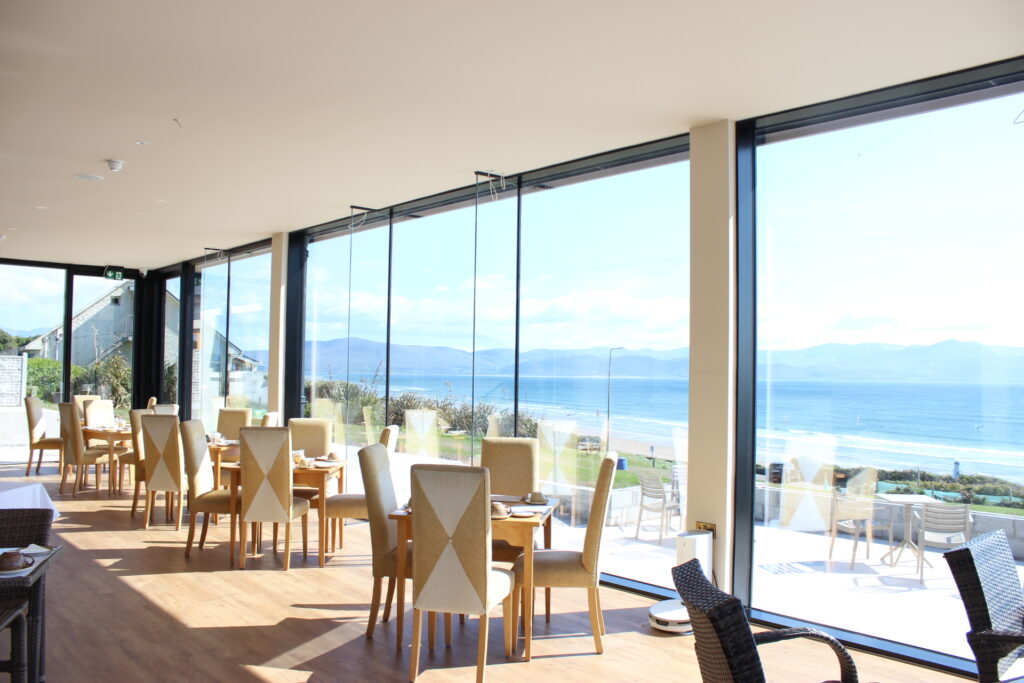 A bright, modern restaurant with large glass windows offers views of the beach and ocean; tables and chairs are set for dining in daylight.