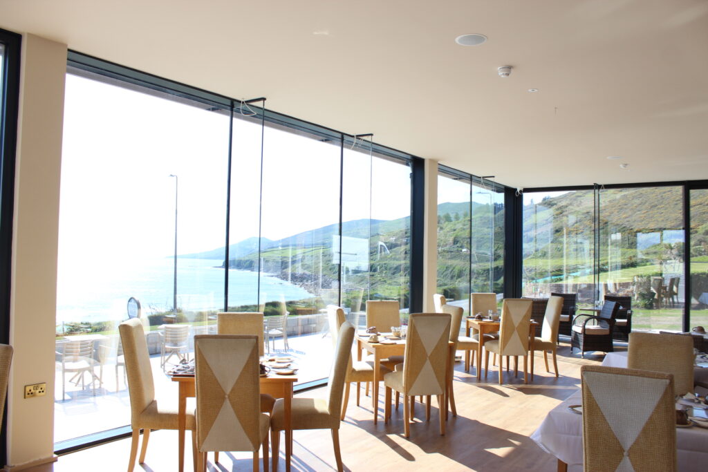 A bright restaurant with large glass windows overlooks a coastal landscape, showing several set tables and wooden chairs inside.