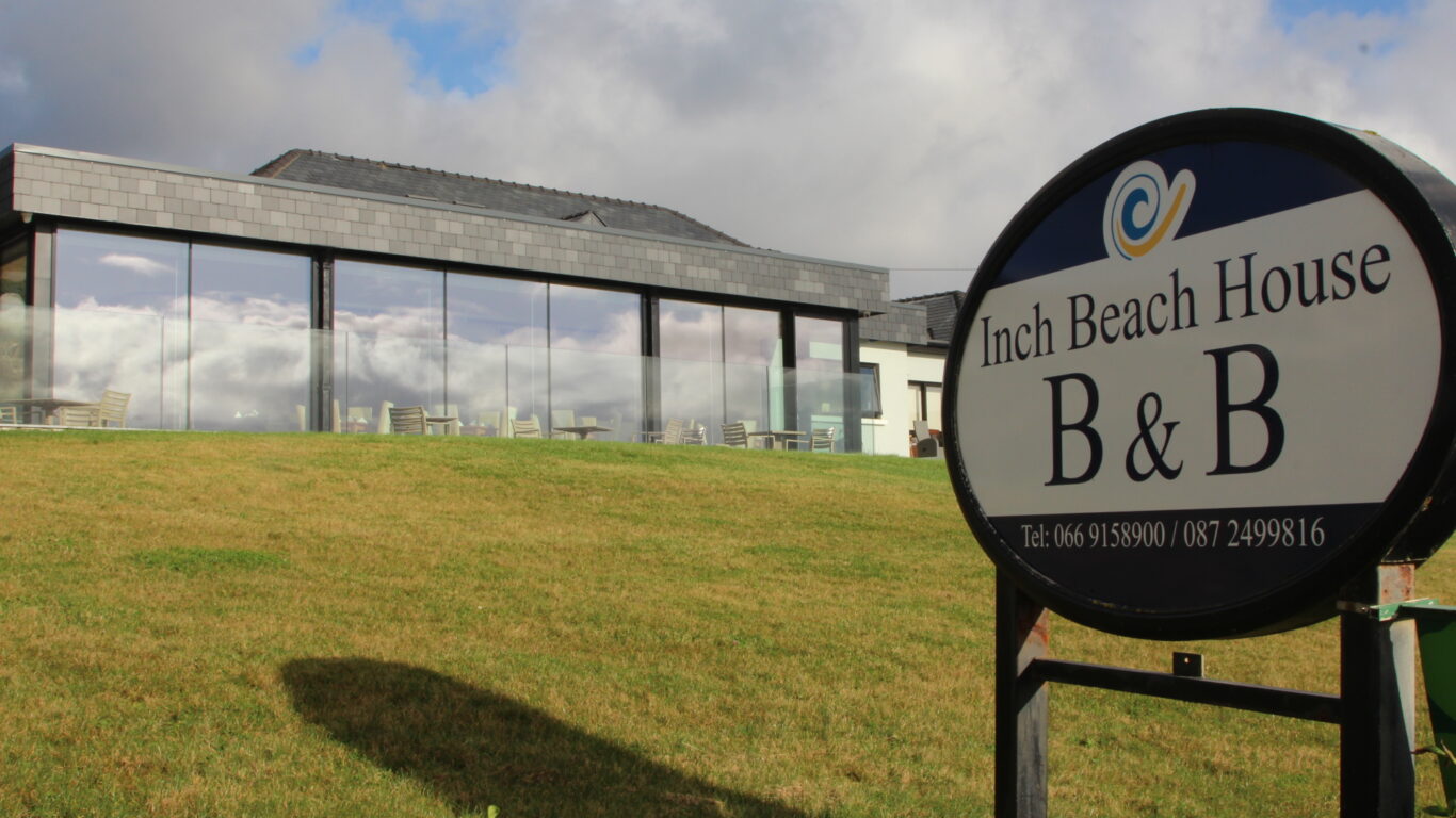 A large sign for Inch Beach House B&B stands on a grassy lawn in front of a modern building with large glass windows under a partly cloudy sky.